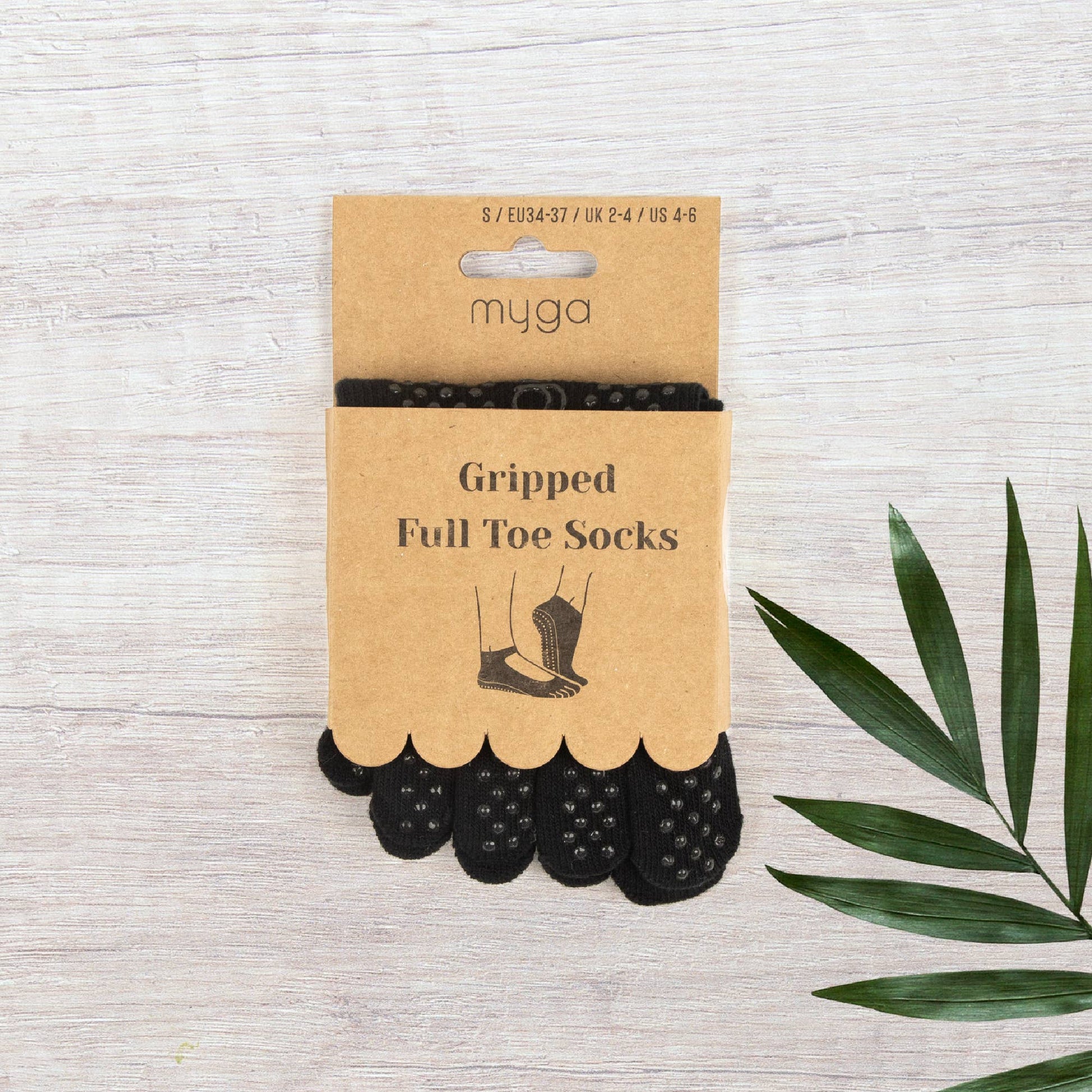Retail packaging for Myga Gripped Full Toe Socks in black. The brown card packaging displays the size information and features a window showing the non-slip grip dots on the sole of the socks. The product is placed on a light wooden background next to a green palm leaf.