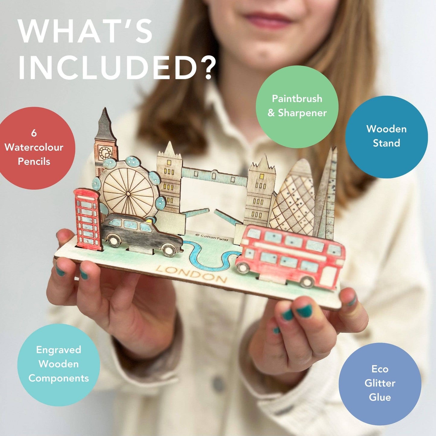 Marketing graphic titled 'WHAT'S INCLUDED?' for the DIY London Landmarks kit. The image highlights the contents over the finished, hand-colored model: 6 Watercolour Pencils, Paintbrush & Sharpener, Engraved Wooden Components (landmarks), a Wooden Stand, and Eco Glitter Glue. A child holds the finished London 3D model in the background.