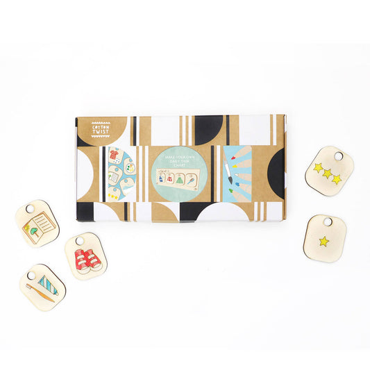 Packaging for the DIY Daily Task Chart or Chore Board craft kit. The product box is surrounded by several hand-colored wooden tokens, including an image of a toothbrush and a sweater, along with two reward tokens featuring stars.