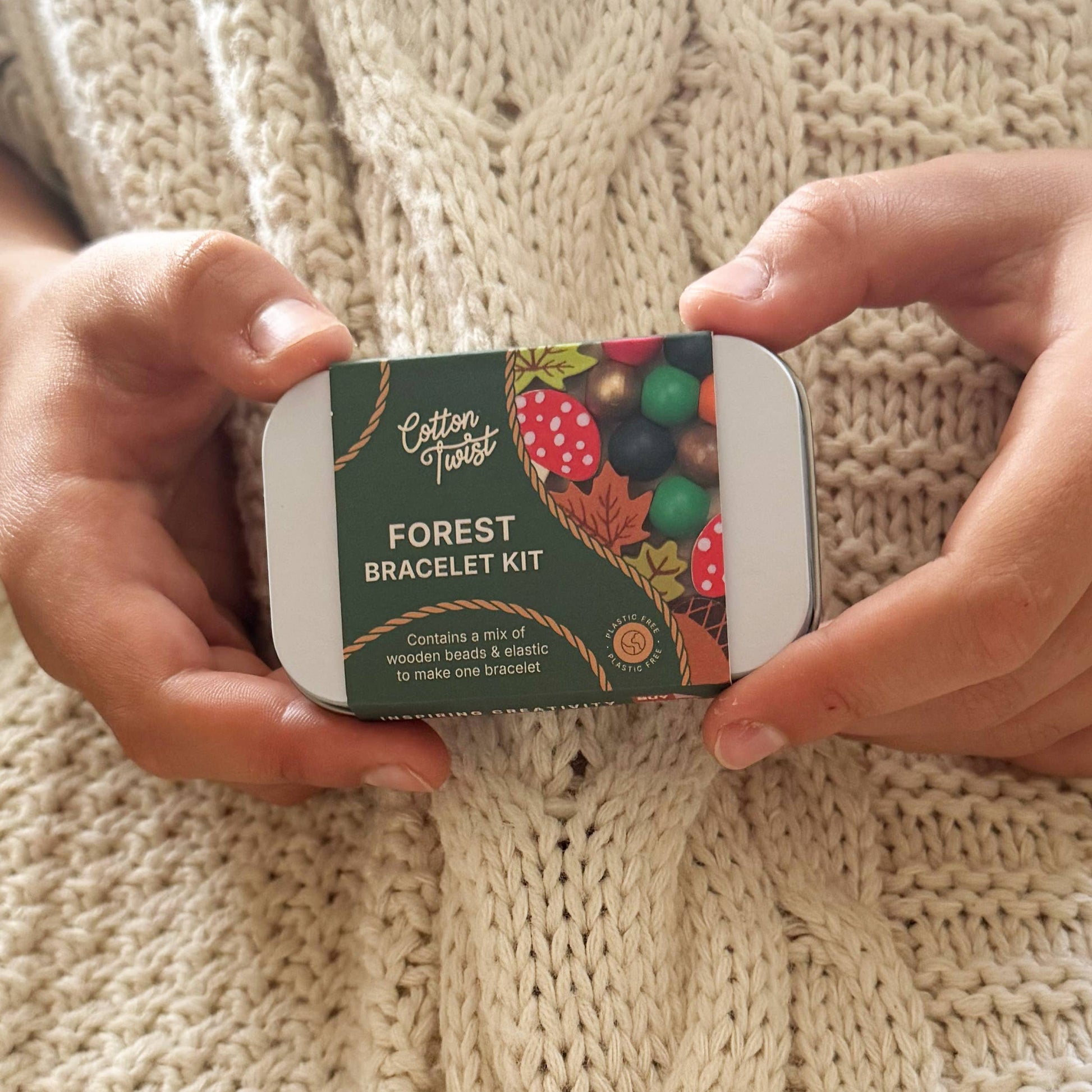 Close-up of the small metal tin packaging for the 'Cotton Twist Forest Bracelet Kit.' The tin is held by a person wearing a cozy, cream-colored cable-knit sweater, emphasizing the autumn and winter craft appeal.