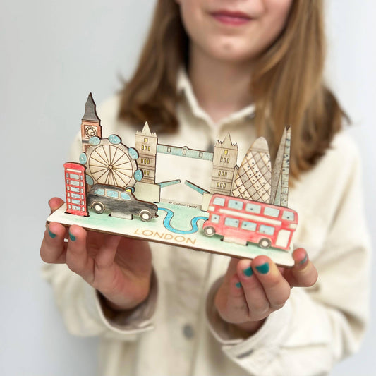 A child holds the completed, hand-colored wooden 3D London scene model. The finished craft features iconic landmarks like the Tower Bridge, Big Ben, the London Eye Ferris wheel, a double-decker bus, a black taxi, and a red telephone booth on a base labeled 'LONDON.'