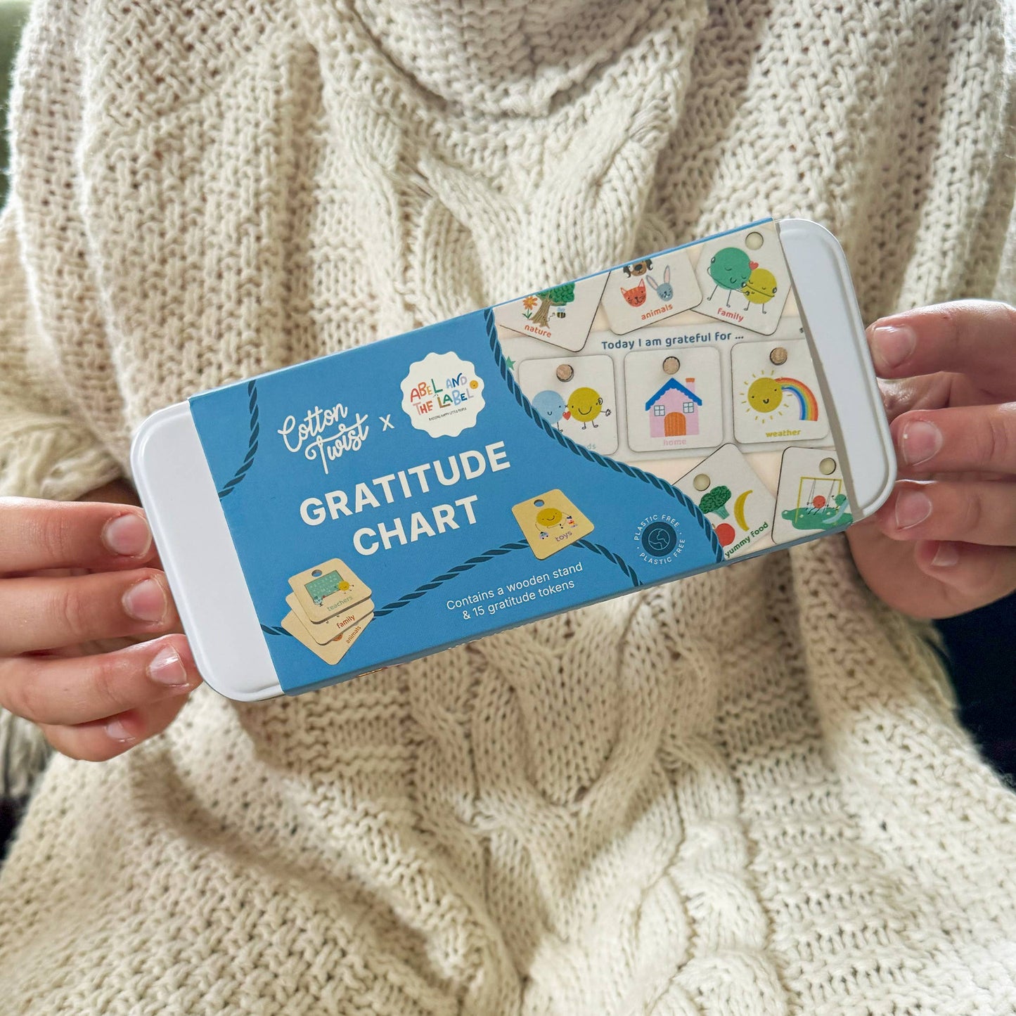 A person wearing a cozy, cream-colored cable-knit sweater holds the blue metal tin packaging for the Cotton Twist Gratitude Chart kit. The tin displays several example gratitude tokens, including a house, rainbow, and tree, highlighting the product's use for promoting thankfulness.