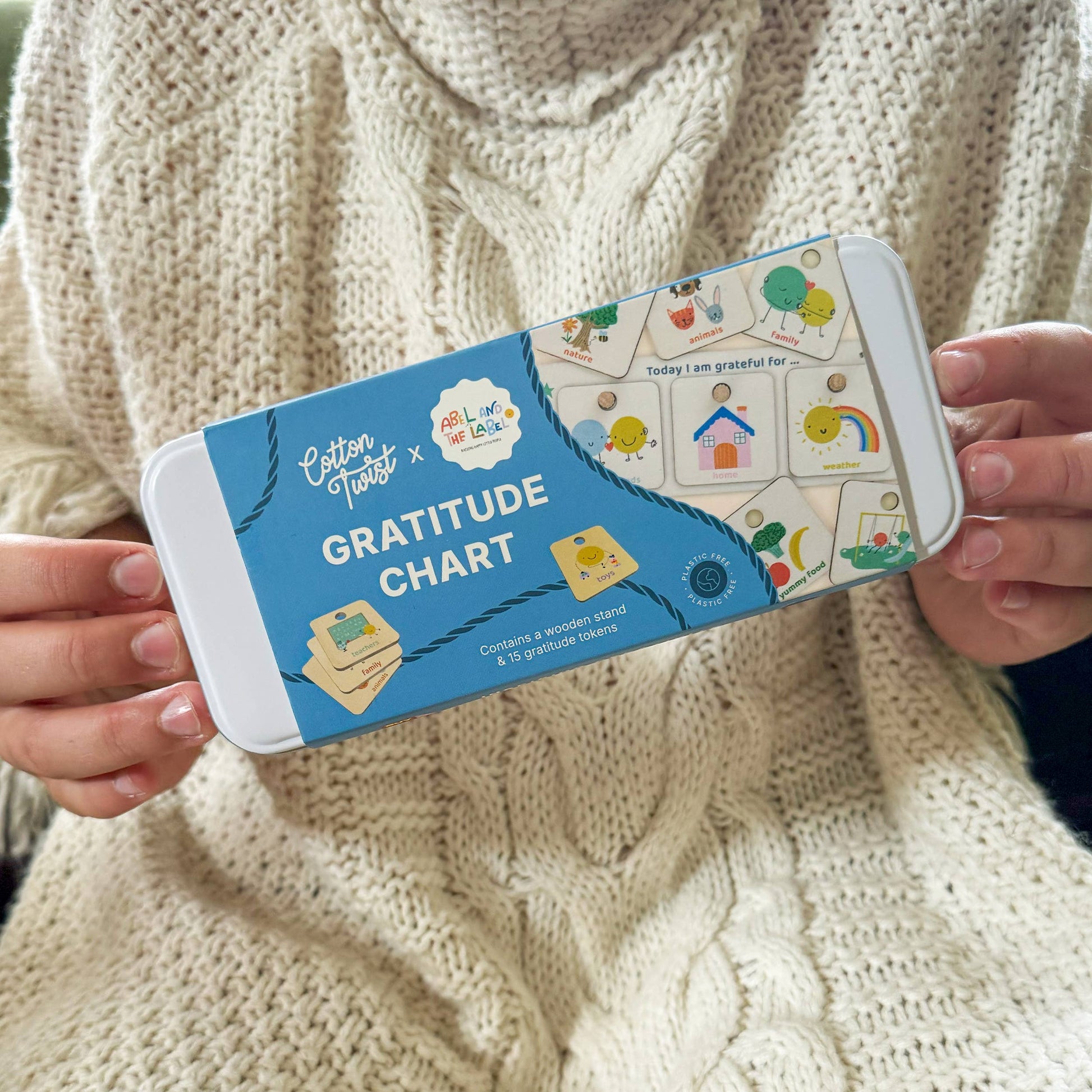 A person wearing a cozy, cream-colored cable-knit sweater holds the blue metal tin packaging for the Cotton Twist Gratitude Chart kit. The tin displays several example gratitude tokens, including a house, rainbow, and tree, highlighting the product's use for promoting thankfulness.