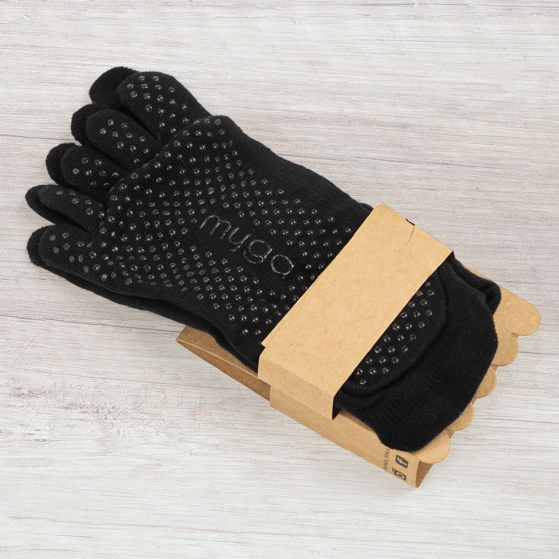 **Close-up of a pair of black full-toe grip socks by Myga, folded with a plain brown cardboard band. The soles clearly show the non-slip dots, highlighting the grip feature for yoga or Pilates practice.