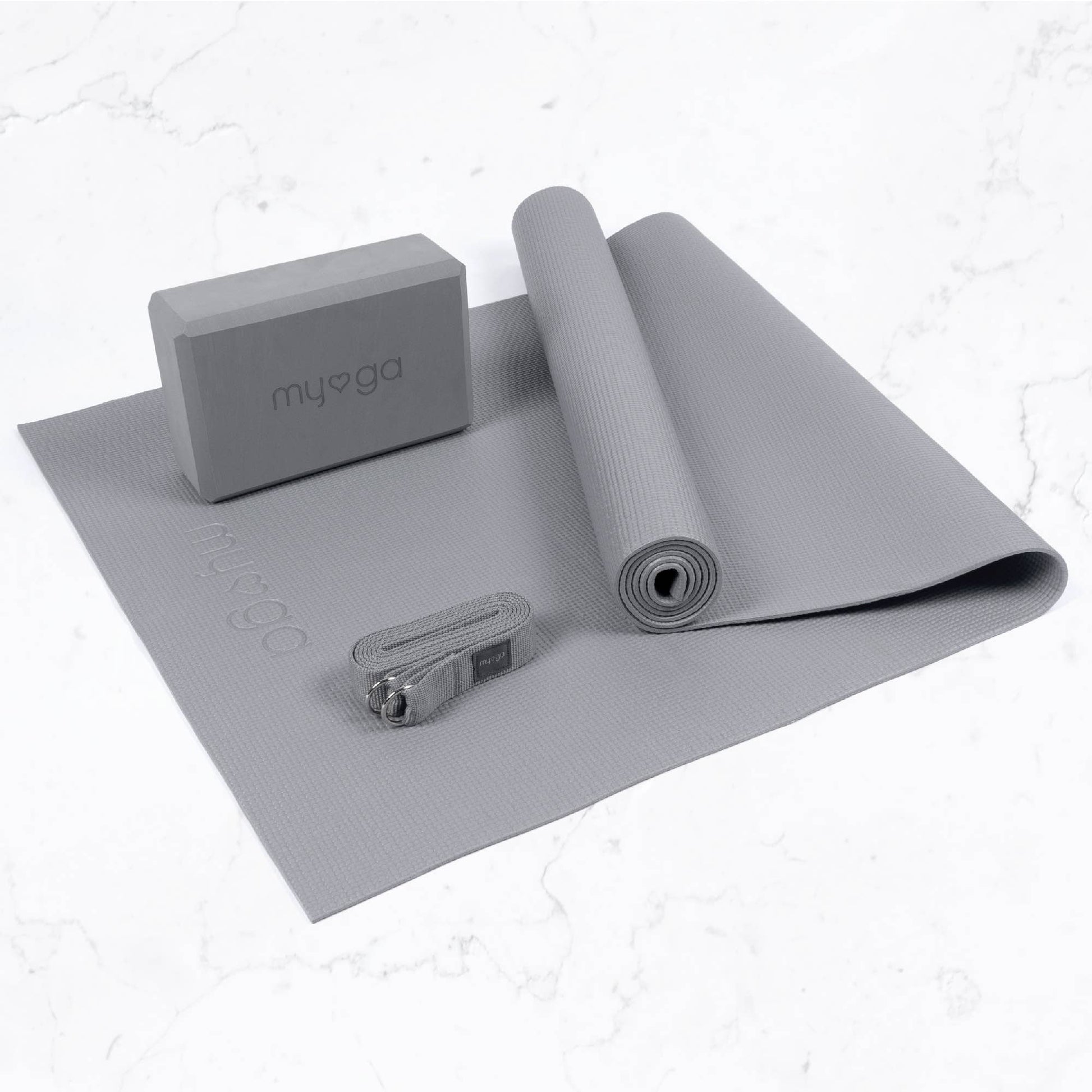 The Myga 3-piece Yoga Set in light grey, featuring a slightly rolled yoga mat, a foam yoga block with the Myga logo, and a yoga strap. The complete set of accessories is displayed on a marble-style background.