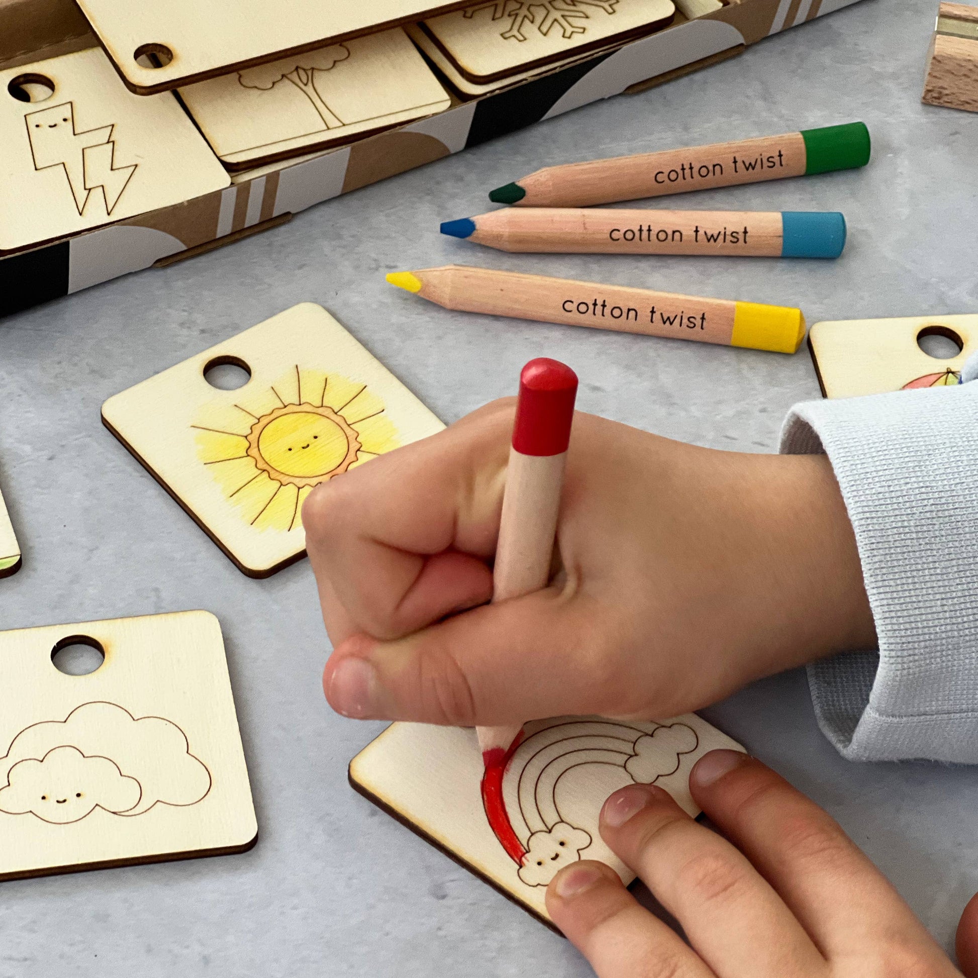 Extreme close-up of a child's hands coloring a wooden rainbow tag with a red wooden pencil from the Cotton Twist craft kit. Uncolored tokens, including a sun and cloud, and two other branded pencils are visible nearby, highlighting the hands-on nature of the DIY weather chart activity.