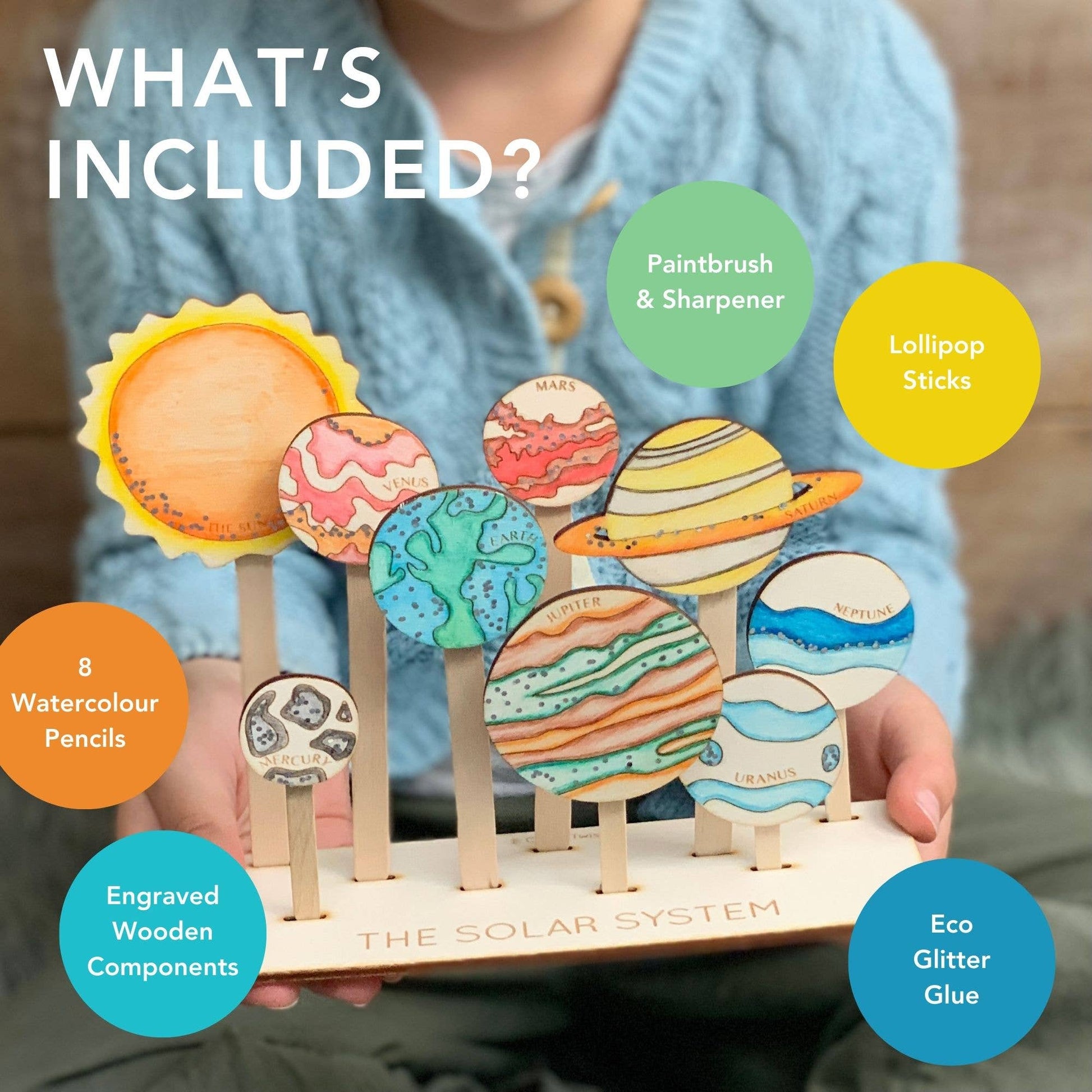 Marketing graphic titled 'WHAT'S INCLUDED?' for the DIY Solar System craft kit. The image highlights the contents over the finished display: 8 Watercolour Pencils, Engraved Wooden Components (planets/sun), Paintbrush & Sharpener, Lollipop Sticks, and Eco Glitter Glue. A child holds the finished, hand-colored wooden solar system display.