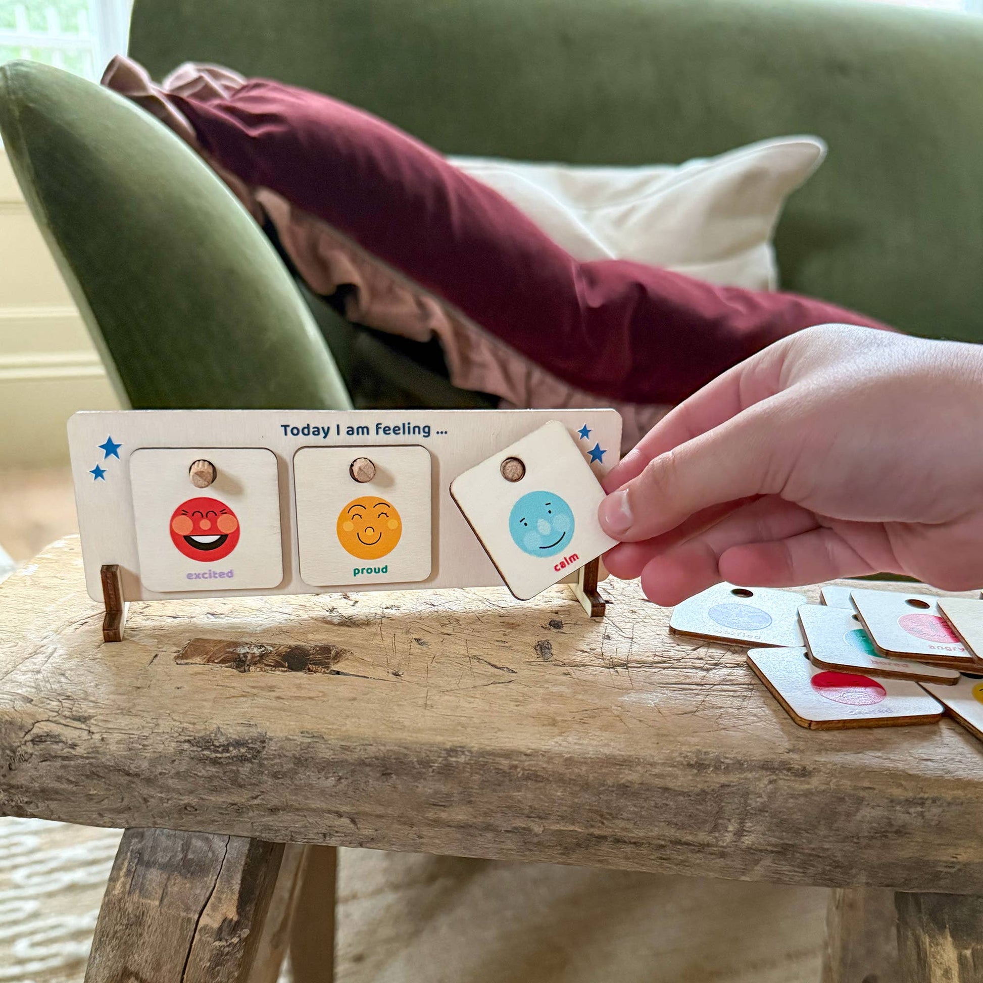 A hand places the 'calm' tile onto the wooden 'Today I am feeling' chart. The chart currently displays 'excited' and 'proud' tiles. The wooden feelings chart is sitting on a rustic wooden table with a soft green velvet couch in the blurred background, highlighting the product in a home setting.