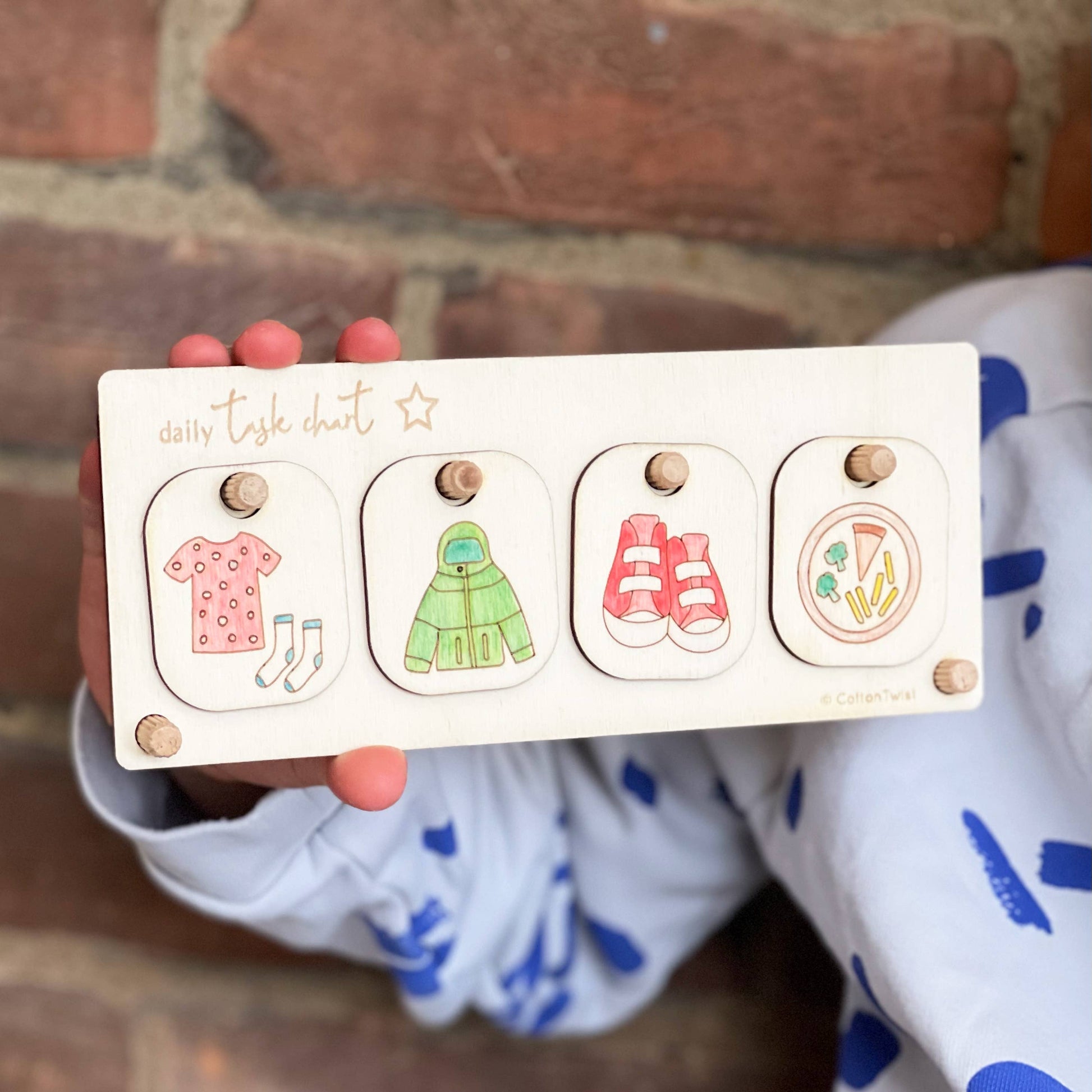 Close-up of a child's hands holding the completed, hand-colored wooden Daily Task Chart against a brick background. The chart displays four chore tokens: getting dressed (dress and socks), putting on a jacket, putting on shoes, and eating vegetables. This shows the product used as a visual routine aid.