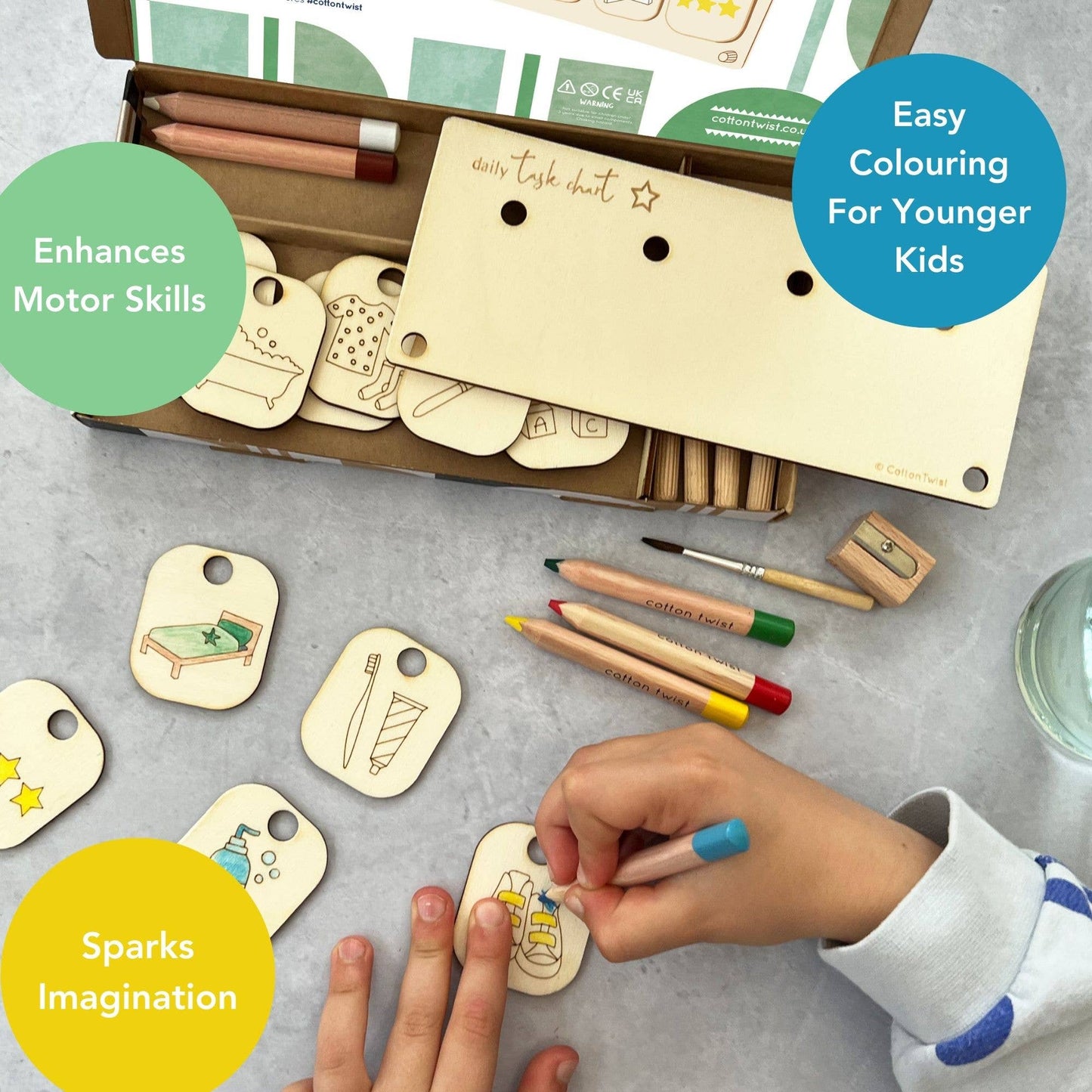 Marketing graphic showing a child coloring a chore token from the DIY Daily Task Chart kit, highlighting the developmental benefits: Enhances Motor Skills, Easy Colouring For Younger Kids, and Sparks Imagination. The open kit box, wooden stand, and other uncolored chore tokens (like making the bed and brushing teeth) are shown in the background.